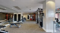 This fitness center offers a comprehensive, thoughtfully organized workout facility spread across a single open floor. The space is finished with neutral carpet flooring, white walls, and recessed lighting throughout, creating a clean and functional environment. Ceiling fans are positioned along the cardio zone, which lines a wall of large windows that invite natural light and outdoor views, providing an energizing atmosphere during workouts.

The cardio section features a row of ellipticals, stair climbers, and treadmills positioned to take advantage of the natural light from the expansive windows. Full-length mirrors are strategically placed throughout the space, enhancing the sense of openness. A dedicated strength training area houses a multi-station cable machine, adjustable benches, and a well-stocked dumbbell rack.

A distinct functional training zone is anchored by a bold orange-accented multi-use rig equipped with TRX straps, medicine balls, kettlebells, battle ropes, and stability balls. A heavy punching bag adds a boxing element to this area. A water fountain is conveniently located near the entrance, and motivational wall signage adds character to the space. The facility is well-ventilated and efficiently laid out to support a wide range of fitness activities.