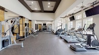 This fitness center offers a generously proportioned, single-floor workout space designed with both functionality and aesthetic appeal in mind. The facility is distinguished by its soaring ceilings accented with dark-trimmed coffered detailing, recessed lighting, and multiple ceiling fans that maintain comfortable airflow throughout the space.

The main workout area is lined with large windows along one wall, flooding the interior with natural light and providing outdoor views. A robust selection of cardio equipment, including treadmills and elliptical machines, is arranged along the windowed wall, each station offering a pleasant sightline. Multiple wall-mounted televisions are positioned throughout for entertainment during workouts.

The strength training section features a comprehensive range of cable and resistance machines, a dumbbell rack with an organized free-weight selection, and additional benches and functional training equipment. Large mirrors are strategically placed to maximize the sense of space and allow users to monitor their form.

A separate entry area showcases striking stone-clad columns and tile flooring, lending an upscale feel to the space. An additional room visible through interior doorways accommodates cycling equipment. The durable rubber flooring throughout the main gym area provides both safety and comfort, completing a well-appointed and inviting fitness environment.