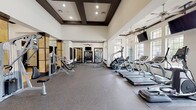 This fitness center offers a generously proportioned, single-floor workout space designed with both functionality and aesthetic appeal in mind. The facility is distinguished by its soaring ceilings accented with dark-trimmed coffered detailing, recessed lighting, and multiple ceiling fans that maintain comfortable airflow throughout the space.

The main workout area is lined with large windows along one wall, flooding the interior with natural light and providing outdoor views. A robust selection of cardio equipment, including treadmills and elliptical machines, is arranged along the windowed wall, each station offering a pleasant sightline. Multiple wall-mounted televisions are positioned throughout for entertainment during workouts.

The strength training section features a comprehensive range of cable and resistance machines, a dumbbell rack with an organized free-weight selection, and additional benches and functional training equipment. Large mirrors are strategically placed to maximize the sense of space and allow users to monitor their form.

A separate entry area showcases striking stone-clad columns and tile flooring, lending an upscale feel to the space. An additional room visible through interior doorways accommodates cycling equipment. The durable rubber flooring throughout the main gym area provides both safety and comfort, completing a well-appointed and inviting fitness environment.
