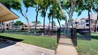 This community dog park offers a spacious, thoughtfully designed outdoor retreat for pets and their owners. The park is fully enclosed by sturdy black metal fencing and features a double-gated entry, providing a secure environment where dogs can roam freely. Lush, well-kept green grass covers the majority of the grounds, offering ample room for exercise and play. Mature trees are distributed throughout the park, providing generous natural shade on sunny days. A dedicated area with mulch ground cover near the entrance serves as a gathering or play zone, complete with benches for owners to relax while their pets enjoy the space. Waste stations are conveniently placed within the park to promote cleanliness. Posted signage at the entrance outlines park rules and hours of operation, indicating the park is open from 6:00 AM to 11:00 PM. The park is set within a larger residential community, surrounded by attractive multi-story buildings that frame the open green space. A covered pavilion structure is visible nearby, adding to the array of outdoor amenities available to residents. The overall ambiance is bright, open, and welcoming, making this dog park a standout community feature.