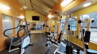 This single-floor fitness center offers a comprehensive workout environment designed to support a wide range of exercise routines. The space is characterized by striking vaulted ceilings with exposed beam detailing and warm yellow-toned wood panel walls, creating an inviting and energizing atmosphere. The open layout accommodates a solid selection of exercise equipment, including cardio machines such as ellipticals and stationary bikes, as well as strength training stations featuring a multi-function cable machine and additional resistance equipment. A wall-mounted flat-screen television provides entertainment during workouts. Full-length mirrors line one wall, enhancing the sense of openness and allowing users to monitor their form while exercising. Large windows along the side wall invite natural light into the space and include a window air conditioning unit, ensuring year-round comfort. The dark rubber flooring is both durable and practical, providing excellent traction and cushioning underfoot. Overall, this fitness center combines functional design with a welcoming aesthetic, making it a well-rounded facility suited for a variety of fitness goals.