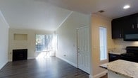 This single-floor residence offers a welcoming and well-lit living environment defined by clean lines and thoughtful architectural details. The living area immediately impresses with its vaulted ceiling, which lends a sense of openness and volume to the space. Crown molding runs throughout, adding a refined finishing touch to the warm, neutral-toned walls.

Rich, dark hardwood-style flooring extends across the main living area, providing a stylish and durable foundation that contrasts beautifully with the bright, white trim and baseboards. Large windows dressed with vertical blinds invite an abundance of natural light, creating an airy and cheerful atmosphere while offering views of the surrounding greenery outdoors.

A standout feature of the living room is the classic fireplace, framed in sleek black, which serves as a natural focal point. Above it, a recessed niche in the wall provides a charming display area. The front entry is flanked by a sidelight window, enhancing the sense of openness upon arrival. A textured ceiling in the entryway transitions to the smooth vaulted ceiling of the main living space, adding architectural interest. Overall, the space presents a comfortable and inviting environment with character-rich details throughout.