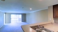 This single-floor residence offers a clean, open-concept layout that seamlessly connects the kitchen and living areas, creating a functional and cohesive living environment. The kitchen is equipped with a double-basin stainless steel sink set into a granite-style countertop, providing ample workspace for meal preparation. The counters wrap around in a curved design, offering a natural partition that defines the kitchen space without fully closing it off from the rest of the home.

The adjoining living area is finished with plush wall-to-wall carpeting in a neutral tone, lending warmth and comfort underfoot. Crown molding runs along the perimeter of the ceiling, adding a refined architectural detail to the space. Recessed lighting provides focused illumination, complementing the natural light that pours in through a large sliding glass door fitted with vertical blinds. This door opens to a private balcony, extending the living space outdoors and allowing for excellent ventilation and natural brightness throughout. The walls are finished in a soft, neutral beige palette, contributing to an overall atmosphere that feels calm, clean, and move-in ready.