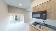 This single-floor studio unit presents a thoughtfully designed open-concept layout finished with light hardwood-style flooring and neutral gray-toned walls throughout. The space is unified by clean lines and a consistent modern aesthetic that creates a bright, functional environment.

The kitchen is compactly arranged yet fully equipped, featuring natural wood-grain cabinetry with matte black hardware, a gray stone-look countertop, a stainless undermount sink with a black faucet, an electric cooktop, an over-the-range microwave, and a compact refrigerator. Recessed lighting above ensures the kitchen remains well-lit.

The main living area benefits from generous natural light, provided by a large picture window and an additional side window, both framing views of the surrounding neighborhood. The vaulted ceiling detail above the living zone adds visual depth and an elevated sense of openness to what might otherwise be a compact space.

A dedicated bathroom features wood-tone cabinetry, a light stone countertop, wood-style flooring, matte black accessories, and clean white walls. An electrical panel is conveniently recessed into the wall near the kitchen entry. The unit's overall design prioritizes practicality and modern simplicity throughout.