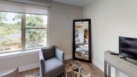 Located at 801 Kenilworth Ave in Charlotte, NC, this single-floor unit offers a thoughtfully designed open-concept layout with modern finishes and a neutral color palette throughout. Warm hardwood-style flooring runs through the main living areas, complementing the soft gray and white tones that define the overall aesthetic.

The living area is bright and inviting, benefiting from large windows that frame views of the surrounding greenery and allow generous natural light to fill the space. A full-length mirror and cozy seating area add both function and style to this comfortable room.

The kitchen is well-appointed with crisp white cabinetry, a white subway tile backsplash, stainless steel appliances, and dark granite countertops. A built-in bar-height island with black stools serves as a casual dining and gathering space, positioned conveniently adjacent to a large window overlooking the outdoors.

The bedroom offers a calm retreat with plush carpeting, soft neutral walls, and warm ambient lighting. Thoughtful décor accents contribute to the room's relaxed, restful atmosphere.

The bathroom features an elongated vanity with dark granite countertops, white cabinetry, undermount sinks, and light tile flooring, creating a clean, spa-like environment. The overall design of this space balances modern functionality with warm, welcoming details.