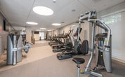 This fitness center is thoughtfully designed to support a wide range of exercise routines within a clean, modern environment. The space is organized into distinct functional zones, allowing users to move seamlessly between cardio, strength, and functional training areas.

The cardio section features a row of Star Trac treadmills alongside elliptical machines and upright bikes, all equipped with individual digital displays. Windows along the walls allow natural light to filter in, contributing to an open and energizing atmosphere. Circular flush-mounted light fixtures in the dropped tile ceiling provide consistent, even illumination throughout.

The strength training area includes a multi-station cable machine, a Hoist functional trainer, and a dedicated free weight zone stocked with dumbbells, kettlebells, and weight plates on organized racks. A flat-screen TV is mounted on the wall for added convenience.

A separate functional training zone offers open floor space with plyo boxes, a heavy bag, resistance bands, and TRX-style equipment, ideal for dynamic or circuit-based workouts. Glass partition walls and doors connect sections of the space, maintaining visual openness while defining separate training areas. Neutral-toned carpet flooring and light grey walls complete the clean, professional aesthetic throughout.
