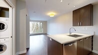 This efficiently laid-out 1-bedroom, 1-bathroom unit offers a thoughtful floor plan that maximizes every square foot across a single level. The open-concept kitchen serves as a focal point, showcasing rich dark-walnut cabinetry, a crisp white quartz peninsula with an undermount sink, and a full suite of stainless steel appliances including a refrigerator, stove, and built-in microwave. Recessed lighting and tile backsplash add refined finishing touches to this culinary space. The living area flows seamlessly from the kitchen, featuring warm hardwood flooring and a large picture window that frames lush, mature tree views, filling the room with natural light and a calming outdoor ambiance. The bedroom is carpeted for comfort and includes sliding mirrored closet doors that enhance the sense of space, along with a dedicated built-in closet organizer. A hallway connects the main living spaces to the full bathroom, which is finished with large-format floor tiles, a deep soaker tub with overhead shower, a wood-paneled vanity with an undermount sink, and a wide mirror. The neutral color palette and clean lines throughout lend the unit a modern, move-in-ready aesthetic.