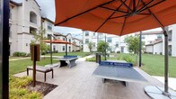 Stella at Riverstone offers a well-appointed amenity complex designed to support an active lifestyle. The fitness center is a standout feature, characterized by soaring vaulted ceilings, large arched windows that flood the space with natural light, and views of the surrounding landscaped grounds. The gym is comprehensively equipped with a row of treadmills and cardio machines positioned along the windowed wall, alongside ellipticals, cable weight machines, free weights, medicine balls, battle ropes, and TRX suspension trainers. Full-length mirrors line one wall, visually expanding the space, while globe pendant lights and ceiling fans add to the comfortable atmosphere. An adjoining corridor features decorative laser-cut barn doors with a geometric floral pattern, built-in cubbies, and access to additional fitness studio space, suggesting a dedicated area for group classes or cycling. Outdoors, a spacious paved courtyard hosts multiple concrete ping-pong tables branded with the Stella at Riverstone name, shaded by large orange market umbrellas. String lights overhead and manicured green lawns surrounded by contemporary white residential buildings complete the inviting outdoor environment.
