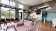 This well-appointed single-floor community space offers a thoughtfully designed layout that blends comfort, functionality, and modern style. The welcoming entrance lobby features rich hardwood-style flooring, tall stone-clad accent walls, a wall-mounted fireplace, and vibrant branded signage that sets an inviting tone from the moment you step inside. Bold pops of red, teal, and charcoal carry throughout the space, lending a lively yet cohesive aesthetic.

The expansive main lounge area is defined by soaring double-height ceilings lined with oversized windows that flood the room with natural light and offer open views of the surrounding greenery. Patterned upholstered sofas and armchairs are arranged around a central gathering area, complemented by a shuffleboard table and a communal dining table with chairs. Exposed ductwork and pendant lighting add an industrial-modern character to the space.

The kitchen and bar area serves as a functional centerpiece, featuring a large white quartz island with bar seating, white shaker cabinetry, subway tile backsplash, stainless steel appliances, and a wall-mounted television. A wood slat ceiling detail above the island adds warmth and architectural interest.

An adjacent seating nook with bold graphic wall treatments and blue accent chairs provides an additional casual retreat, completing this versatile and engaging community environment.