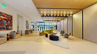 Located in the heart of Miami at 777 Northwest 2nd Avenue, this expansive ground-floor lobby is a masterclass in modern residential design. The grand entrance foyer welcomes residents and guests through floor-to-ceiling glass doors framed in warm wood tones, offering immediate views of the surrounding streetscape and a beautifully paved outdoor courtyard beyond.

The main lounge area commands attention with its soaring ceilings clad in rich wood paneling, punctuated by rows of recessed and track lighting that cast a warm, inviting glow across large-format stone tile flooring. Curated seating arrangements — including olive-green curved sofas, classic Eames lounge chairs, and sculptural ottomans — are anchored by a geometric area rug, creating distinct conversation zones within the generous open floor plan. A sleek gold reception desk serves as a striking focal point near the entrance, complemented by lush potted tropical plants throughout.

A dedicated hallway leads to the well-appointed mail room, lined with banks of dark bronze mailboxes and adorned with abstract wall sculptures and trailing greenery. Nearby, a set of stylish private phone booths provides quiet, enclosed spaces for focused work or calls. A bright conference room features a long hardwood table beneath a statement brass chandelier, wall-sized botanical murals, and expansive windows overlooking the street — ideal for meetings or collaborative use.
