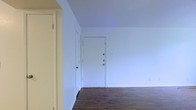 This single-floor space presents a clean and straightforward layout defined by its bright white walls and rich dark hardwood flooring. The open entryway area is immediately welcoming, anchored by a solid white front door fitted with two deadbolt locks, offering both security and a crisp aesthetic. The smooth, white walls throughout create a neutral backdrop that maximizes the sense of openness and allows for versatile interior styling. Multiple doorways are visible from the main area, indicating a practical floor plan with clear transitions between spaces. The dark hardwood floors provide a warm contrast to the light walls, grounding the space with a classic, timeless character. The layout is compact yet functional, making efficient use of available space across the single floor. The overall ambiance is minimal and understated, offering a blank canvas that is well-suited for a range of furnishing and décor choices. The absence of clutter and the clean architectural lines contribute to a sense of calm and order throughout the space.