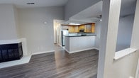 This single-floor residence offers a clean, open-concept layout that seamlessly connects the living area and kitchen, creating a sense of spaciousness and ease of movement throughout. The kitchen is a functional highlight of the home, featuring warm honey-toned wood cabinetry, dark countertops, and stainless steel appliances including a refrigerator and microwave. A peninsula-style breakfast bar separates the kitchen from the living space, providing both a casual dining option and an architectural division between the two areas. Recessed and overhead lighting illuminates the kitchen workspace effectively. The living area is generous in scale, bathed in a neutral gray palette that lends a modern, understated aesthetic. Durable hardwood-style flooring in a cool gray-brown tone runs consistently throughout, tying the spaces together cohesively. Vaulted or elevated ceilings in the main living zone add verticality and a sense of openness. A hallway visible beyond the kitchen leads to additional areas of the home. The overall ambiance is contemporary and move-in ready, with clean lines, neutral tones, and practical finishes that offer a versatile backdrop for a variety of interior styles.
