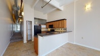 Located at 22 S Saint Clair St in the heart of downtown Dayton, Ohio, this single-floor loft-style unit blends industrial character with modern residential comfort. Dramatic soaring ceilings with exposed structural beams and visible ductwork give the space a distinctive urban aesthetic throughout. Large-format tile flooring runs through the main living and kitchen areas, reflecting abundant natural light that pours in through expansive floor-to-ceiling windows fitted with vertical blinds.

The open-concept kitchen is a functional centerpiece, featuring warm honey-toned wood cabinetry with crown molding, dark granite-style countertops, a decorative mosaic tile backsplash with under-cabinet lighting, and a full suite of black appliances including a range, over-the-range microwave, and refrigerator. A peninsula with a matching countertop provides additional prep and casual dining space, opening seamlessly to the adjacent living area.

The bedroom is generously sized with plush carpeting and its own floor-to-ceiling window wall, offering urban views and excellent natural light. A walk-in closet with built-in shelving and hanging organizers provides ample storage. Two full bathrooms, each with a tub/shower combo and tile flooring, serve the space. The unit's elongated layout, as seen from the dollhouse view, offers a practical and well-organized flow throughout.