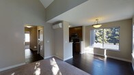This single-floor residence offers a thoughtfully designed open-concept layout that seamlessly connects the main living areas. The space is anchored by rich, dark hardwood flooring that extends throughout the kitchen and living areas, contrasting with plush carpet in the lounge zone. Soaring vaulted ceilings lend an airy, expansive feel to the interior, enhancing the sense of openness throughout.

The kitchen is efficiently arranged with dark wood cabinetry, stainless steel appliances including a built-in microwave, and a compact peninsula that defines the space without interrupting the flow of the floor plan. A wall-mounted mini-split HVAC unit provides targeted climate control for year-round comfort.

Natural light pours in through a large picture window, offering pleasant outdoor views of mature trees and filling the living area with warmth and brightness. A pendant light fixture above the dining or entry zone adds a decorative touch while providing functional illumination.

The entry hallway, visible from the main living space, leads to additional rooms and features its own natural light source, contributing to the overall bright and welcoming ambiance. Neutral wall tones throughout the space complement the dark flooring and cabinetry, creating a cohesive and modern aesthetic.