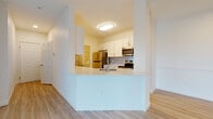 Located at 1104 S Missouri Ave in Clearwater, Florida, this single-floor unit offers a clean, modern interior defined by a neutral color palette and light wood-style flooring that runs consistently throughout the space. The open-concept layout connects the living area to the kitchen, which features a white peninsula with an integrated sink, white shaker-style cabinetry, a tiled backsplash, and a full suite of stainless steel appliances including a refrigerator, range, and microwave. The living area is generously proportioned, with a ceiling fan providing comfort overhead and a tray ceiling detail adding architectural interest. A dedicated laundry area houses a full-size washer and dryer with built-in wire shelving above for additional storage. The bedrooms are bright and well-sized, with large windows fitted with horizontal blinds allowing natural light while maintaining privacy. The bathrooms each include a tiled shower-tub combination with a curved curtain rod and wood-style flooring. A thermostat and closet with wire shelving are visible near the entryway, contributing to the unit's practical and well-organized design.