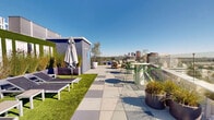 Located atop a building on the iconic Wilshire Boulevard in Beverly Hills, this rooftop terrace offers a sophisticated outdoor experience with sweeping 360-degree panoramic views of the greater Los Angeles skyline, the Hollywood Hills, and surrounding urban landscape. The terrace is thoughtfully designed with a mix of large-format porcelain tile paving and sections of lush artificial turf, creating a dynamic and visually engaging surface throughout the space. Glass perimeter railings ensure unobstructed sightlines while maintaining an open, airy atmosphere under the abundant Southern California sun. Multiple distinct seating zones are distributed across the terrace, including cushioned lounge chairs and chaise lounges ideal for relaxation, as well as teak-framed conversational seating arrangements suited for gatherings. A bar-height table with green metal stools offers a casual dining or socializing option. A white pergola structure provides a shaded retreat, complementing the open sun-drenched areas. Potted ornamental grasses, trees, and sculpted planters add greenery and a resort-like ambiance throughout. Outdoor shower access and utility structures are integrated discreetly into the design. The overall aesthetic is modern and clean, perfectly suited to the prestigious Beverly Hills address.