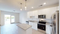 This single-floor residence offers a clean, contemporary design highlighted by an open-concept layout that seamlessly connects the kitchen and living areas. The kitchen is a standout feature, outfitted with crisp white shaker-style cabinetry extending to the ceiling for ample storage, granite countertops, and a full suite of stainless steel appliances including a refrigerator, electric range, built-in microwave, and dishwasher. A central kitchen island with a matching granite surface provides additional prep space and serves as a natural gathering point. Elegant pendant lighting hangs above the island, complementing the recessed ceiling lights throughout. Light-toned hardwood-style flooring flows through the main living areas, adding warmth and continuity to the space. The bedroom features neutral carpeting and windows with blinds that invite natural light and offer views of the surrounding neighborhood. The bathroom is well-appointed with white cabinetry, granite countertops, an undermount sink, a large vanity mirror, and a tall linen tower for added storage. A dedicated in-unit laundry area with a stacked washer and dryer adds everyday convenience. The overall aesthetic is modern and neutral, with a bright, airy ambiance throughout.