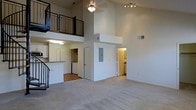 This two-story space is defined by its striking double-height ceilings in the main living area, creating an expansive, open atmosphere. The ground floor features neutral wall-to-wall carpeting that extends throughout the living space, while hard flooring is visible in the adjacent kitchen and utility areas. A staircase along one wall leads to the upper level, where a loft-style balcony with black metal railing overlooks the main floor below — a standout architectural detail that adds both visual interest and a sense of grandeur.

The kitchen area is partially open to the living space, separated by a raised counter that serves as a natural divider. White cabinetry lines the kitchen walls, and a laundry area with side-by-side units is conveniently situated nearby. A utility panel is accessible on the main floor wall. Track lighting on the upper portion of the living area and a ceiling fixture provide artificial illumination throughout. Additional rooms are accessible through doorways visible from the main living space, including what appears to be a bathroom with warm lighting. The overall aesthetic is clean, neutral, and functional.