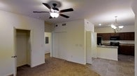 This single-floor space offers a practical and open layout designed for comfortable everyday living. The main living area features wall-to-wall carpeting that adds warmth underfoot, while a ceiling fan with a built-in light fixture provides both air circulation and ambient lighting overhead. The walls are finished in a neutral tone, creating a bright and clean backdrop throughout.

A prominent feature of the main room is the large double-door closet, offering generous storage capacity. Adjacent to the closet, a doorway leads into the kitchen, which transitions to tile flooring and is equipped with dark wood cabinetry and a light-toned countertop, providing a functional contrast to the living space. Recessed lighting in the kitchen adds a focused, practical illumination to the cooking area.

A second doorway from the main room provides additional access, suggesting a connected and efficiently organized floor plan. Vents are visible along the upper walls, indicating a central HVAC system for year-round climate control. The overall layout is straightforward and well-proportioned, making it easy to navigate and furnish. The neutral color palette and consistent finishes throughout give the space a cohesive and move-in-ready appearance.