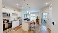 This efficient and thoughtfully designed 1-bedroom, 1-bathroom residence offers a comfortable single-floor layout. The open-concept kitchen and living area serves as the heart of the space, where the kitchen features crisp white shaker cabinetry extending to the ceiling, stainless steel appliances including a refrigerator, range, microwave, and dishwasher, a gray quartz-style countertop, subway tile backsplash, and a central island with seating for two, illuminated by stylish pendant lighting. Light wood-style flooring runs throughout this area, enhancing the bright, airy atmosphere. The living room offers generous natural light through large windows and a sliding glass door that opens onto a private balcony with outdoor views. The bedroom is a calm, carpeted retreat with a large window that draws in natural light, offering a comfortable and quiet space. A dedicated closet provides ample storage. The bathroom is clean and modern, featuring white shaker-style cabinetry, a gray countertop vanity, a large mirror with overhead lighting, and a full tub-shower combination with white tile surround. Neutral wall tones and consistent modern finishes create a cohesive, welcoming ambiance throughout the entire residence.
