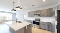 This single-story, 3-bedroom, 2-bathroom home delivers a clean, modern aesthetic with consistent light hardwood flooring and neutral tones throughout. The open-concept living area seamlessly connects the kitchen, dining, and living spaces, creating a bright and functional hub for daily life. The kitchen is a standout feature, showcasing gray shaker-style cabinetry, a crisp white subway tile backsplash, stainless steel appliances including a range and over-the-range microwave, and a central island with a white quartz countertop, undermount sink, and stylish pendant lighting above. The bedrooms are calm and bright, with large windows offering views of surrounding greenery. The primary bedroom includes a dedicated walk-in closet with wire shelving. Five closets total provide generous storage throughout the home. The two bathrooms feature subway tile surrounds — one with a soaking tub and matte black fixtures, and the other with a sleek sliding glass shower enclosure and a backlit mirror. An in-unit laundry area houses a stacked washer and dryer. A utility room rounds out the practical amenities of this well-organized and move-in ready home.
