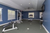 Located at 150 Orchard Brook Dr in Gainesville, Georgia, this single-floor fitness center is thoughtfully designed to support a well-rounded workout experience. The space is defined by its striking deep blue walls, crisp white baseboards, and durable carpet flooring throughout, creating a focused and energizing atmosphere. Recessed lighting is complemented by a ceiling fan, ensuring the room remains comfortable and well-illuminated during exercise sessions. Multiple windows fitted with blinds allow natural light to filter in while maintaining privacy. The fitness area is generously proportioned, offering ample room for a variety of equipment and movement. A treadmill and elliptical machine serve as the primary cardio stations, while a cable/pulley strength training machine and a leg extension/curl bench provide dedicated strength training options. An adjacent room, visible through an open doorway, features hardwood-style flooring and appears to function as an office or lounge area, adding versatility to the overall facility. Safety features including a fire extinguisher and emergency exit signage are present throughout. The layout is open and uncluttered, making the space practical and easy to navigate for users of all fitness levels.