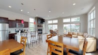 This well-appointed single-floor community clubhouse offers an open-concept design filled with abundant natural light streaming through large multi-pane windows. The layout seamlessly integrates several functional zones, creating a welcoming and versatile gathering space.

The kitchen is fully equipped with stainless steel appliances, including a refrigerator, electric range, and built-in microwave, all complemented by rich dark cabinetry and light granite-style countertops. Striking red pendant lights add a pop of color and character above the kitchen bar area, which features stylish counter-height seating.

Adjacent to the kitchen, a cozy fireplace with a wall-mounted television above serves as a central focal point, anchoring the casual dining and lounge areas. The lounge features a generously sized wraparound leather sectional sofa positioned to take full advantage of the floor-to-ceiling windows, which flood the space with natural light and offer views of the surrounding community.

Multiple dining configurations accommodate groups of varying sizes, from intimate gatherings to larger social events. Light wood-tone flooring runs throughout, enhancing the bright, airy atmosphere.

Large sliding glass doors and oversized windows open directly to an outdoor pool deck furnished with seating and umbrellas, providing a seamless indoor-outdoor connection that makes this space ideal for community gatherings and recreational use.