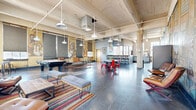 Located at 2400 S Ervay St in Dallas, TX, this single-floor industrial-style space offers an expansive and versatile layout that blends raw architectural character with curated, functional design. The main open area is defined by soaring ceilings with exposed beams, structural columns, and pendant barn lights that cast a warm glow over polished concrete floors. Bold large-scale murals depicting jazz musicians and vibrant urban scenes adorn the walls, lending the space a distinct artistic identity. The lounge is furnished with eclectic seating, colorful area rugs, and a pool table, creating an inviting communal atmosphere. A bar-style counter and dining area with cross-back chairs provide additional gathering space. The gym features vivid teal-painted walls, large industrial windows that flood the room with natural light, and a comprehensive selection of strength and cardio equipment. An adjacent studio room offers additional flexibility for fitness or wellness activities. The bathroom is clean and contemporary, finished with light wood-look flooring and a stylish navy vanity with gold accents. The outdoor patio extends the space further, offering a stamped concrete terrace with string lights overhead, tiered wooden bleacher-style seating, a grill, and open views of the surrounding Dallas skyline.