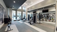 This contemporary fitness center is designed with both functionality and aesthetics in mind. The space features soaring double-height ceilings accented by a combination of dark and light tonal finishes, creating a dynamic and energizing atmosphere. Large floor-to-ceiling windows along the perimeter flood the workout area with natural light while offering views of the surrounding outdoor landscape.

The facility is divided into distinct training zones. The cardio area is lined with a row of treadmills and elliptical machines, each equipped with individual digital displays. The strength training section includes a full complement of cable machines, weight benches, and Smith machines, supported by a wall of floor-to-ceiling mirrors that enhance the sense of space and allow for proper form checks. A dedicated free weights area features a dumbbell rack alongside adjustable benches, stability balls, and spin bikes.

A bold black-and-white athletic mural serves as a striking focal point on one wall, adding a motivational aesthetic element. Patterned carpet tiles in neutral gray tones define the floor throughout, and recessed lighting combined with a ceiling fan ensures comfort during intense workouts. A welcoming entry area with a fitness rules board and a small beverage station provides a polished, professional touch to the overall experience.