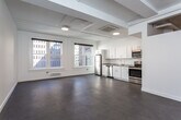This single-floor unit presents a clean, contemporary design characterized by an open-concept layout and a neutral palette throughout. The main living area is expansive and airy, featuring smooth flooring, exposed structural ceiling beams painted white, and ceiling-mounted HVAC units that maintain a streamlined aesthetic. Large windows frame views of the surrounding urban cityscape, drawing in natural light and reinforcing the modern character of the space.

The kitchen runs along one wall and is well-appointed with white cabinetry, granite-style countertops, and a full suite of stainless steel appliances including a refrigerator, dishwasher, electric range, and microwave. The open connection between the kitchen and living area enhances the sense of spaciousness.

The bedroom is a generously sized, carpeted room with its own natural light source and is complemented by a dedicated walk-in closet equipped with built-in shelving, hanging rods, and a drawer unit, offering substantial storage capacity.

The bathroom features a marble-look vanity, a glass-enclosed shower, and durable tile-style flooring. Throughout the unit, the consistent use of white walls, neutral flooring, and minimal detailing creates a cohesive, functional living environment.
