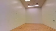 This single-level space is a purpose-built racquetball court designed to meet the demands of both casual players and serious competitors. The court features a classic hardwood floor finished in a warm natural tone, with clearly marked court lines that define the playing boundaries. The smooth, seamless white walls on all sides are purpose-designed to ensure consistent ball rebound, a hallmark of a properly constructed racquetball environment. The high ceiling provides ample vertical clearance, accommodating the full range of overhead shots and lob plays integral to the game. Overhead fluorescent panel lighting is evenly distributed across the ceiling, ensuring uniform illumination throughout the court with minimal shadowing — an important feature for tracking a fast-moving ball. Ventilation grilles are integrated into the ceiling, supporting comfortable air circulation during intense play. The clean, minimalist design of the space keeps the focus entirely on athletic performance, with no visual distractions. Overall, this well-maintained racquetball court offers a functional and regulation-ready environment suited for individual practice sessions, competitive matches, or structured group programming.