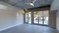 This single-floor space showcases a compelling blend of industrial and contemporary design elements. The exposed concrete ceiling is a defining architectural feature, displaying raw texture and natural variation that adds character and depth to the interior. A ceiling fan with a warm brass fixture is centrally mounted, providing both function and a subtle decorative touch.

The flooring consists of smooth, polished concrete that complements the ceiling and contributes to the space's cohesive, modern industrial aesthetic. The neutral-toned walls provide a clean contrast against the raw concrete surfaces, creating a balanced visual environment.

Natural light is a prominent feature, courtesy of a large wall of floor-to-ceiling glass sliding and French-style doors that span the exterior-facing wall. These doors offer direct views of a lush, landscaped outdoor area featuring palm trees and manicured plantings, seamlessly connecting the interior to the outdoors. Roller shades are fitted to each door panel, offering adjustable light control and privacy.

The open layout of the space provides versatility, making it well-suited for a variety of uses. The combination of raw materials, generous natural light, and indoor-outdoor connectivity gives this space a distinctive, airy ambiance with strong architectural presence.