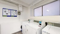 Located at 855 Jervis St in Vancouver's West End, this shared laundry facility provides a clean, well-organized space designed for practical, everyday use. The room is finished with light-toned tile flooring and crisp white walls, creating a bright and functional environment.

The laundry area is equipped with multiple Huebsch top-load washers and dryers, all operating via Coinamatic coin-payment systems. Machines are arranged along the walls, allowing for efficient use of the available floor space. Instructional signage and usage guidelines are posted prominently near the machines for user reference.

A dedicated folding station features a smooth, speckled white countertop with white shaker-style cabinetry below and open shelving above, offering convenient storage and a surface for sorting and folding laundry. A security camera is mounted near the ceiling, and a window allows natural light into the space. A framed aerial photograph of Vancouver adds a modest decorative touch to the otherwise utilitarian room. The overall atmosphere is clean, practical, and well-maintained, serving the needs of building residents efficiently.