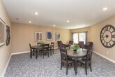 Located on Iris Drive in Conyers, Georgia, this single-floor space offers a warm and inviting atmosphere designed for comfortable gathering and relaxed conversation. The interior is finished with neutral, earthy tones on the walls, creating a calm and approachable ambiance throughout.

The sitting area is furnished with a set of plush, deep-toned wingback armchairs arranged around a glass-top coffee table, providing an ideal setting for casual interaction or quiet reflection. Natural light filters in through multiple windows dressed with horizontal blinds and draped curtains, lending the space a bright and airy quality. A large framed abstract artwork adds a thoughtful decorative accent to the sitting area walls.

Adjacent to the seating area, a carpeted dining nook features a round wooden table accompanied by dark-finished chairs, creating an intimate space for small group meals or meetings. The walls are adorned with distinctive textured wall art panels and a framed print, contributing to the space's curated, welcoming character.

Patterned carpeting flows throughout, adding softness underfoot and visual warmth. Recessed lighting and ceiling-mounted vents ensure consistent comfort across the space. The overall design balances functionality with a homey aesthetic, making this a versatile and comfortable environment for residents or visitors alike.