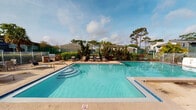 This outdoor recreational area centers around a generously sized swimming pool with crystal-clear turquoise water, framed by a wide concrete sundeck designed for relaxation and socializing. The pool features a gradual entry staircase on one end and a dedicated shallow section clearly marked for safety, making it accessible for a range of swimmers. Surrounding the pool, an ample deck is furnished with multiple lounge chairs and side tables, providing comfortable spaces for sunbathing and leisure throughout the day. A built-in outdoor grill station is conveniently positioned poolside, ideal for gatherings and outdoor dining. The area is enclosed by a white metal fence, ensuring privacy and security, while lush landscaping including mature pine trees, neatly trimmed shrubs, and decorative plantings create a natural, park-like atmosphere. Tennis courts are visible just beyond the perimeter, adding to the range of recreational amenities available on the grounds. A single-story support structure adjacent to the pool deck provides additional utility to the space. The open sky above and the canopy of surrounding trees contribute to a bright, airy ambiance that is both inviting and functional for outdoor community activities.