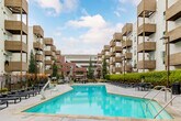 Located at 868 S Fifth St in San Jose, California, this outdoor amenity space is a well-appointed resort-style pool courtyard designed for relaxation and recreation. The centerpiece is a generously sized swimming pool with striking turquoise water, complemented by sleek gold-toned handrails and a separate spa/hot tub area positioned at one end. The pool deck is finished in smooth concrete and is lined with multiple modern chaise lounges and outdoor dining tables with seating, providing ample space for sunbathing and socializing. A distinctive woven pod chair nestled against a textured brick accent wall adds a unique lounging option to the space. The courtyard is framed by a lush, manicured hedge wall interspersed with dramatic agave and succulent plantings, creating a verdant and low-maintenance landscape. Wrought iron fencing defines the perimeter, offering both security and an open, airy feel. The surrounding multi-story building features a contemporary architectural design, with bold yellow and white exterior panels and wood-accented cantilevered balconies that overlook the courtyard. The overall ambiance is modern and urban yet resort-like, offering a well-designed outdoor retreat in the heart of San Jose.