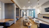 This expansive single-floor amenity space is designed with a modern yet warm aesthetic, blending natural materials with contemporary furnishings throughout. The layout flows from a reception area featuring a light wood desk, a wall-mounted fireplace, and an accent wall with botanical wallpaper, creating an inviting first impression. Recessed and track lighting illuminate the white ceilings, while exposed wood beam accents and large windows allow natural light to filter throughout.

The main lounge zones are furnished with plush, rounded sofas in neutral and amber tones, complemented by organic-shaped coffee tables and woven area rugs. Cozy semi-private booth seating with navy upholstery and pendant lighting offers intimate gathering spots along the corridor. A separate lounge features floor-to-ceiling sheer drapes, a statement chandelier, and tufted leather sectionals.

Dedicated work spaces include glass-enclosed private work-from-home pods and a conference room with a long walnut table and carpeted flooring. Black-framed sliding barn doors with frosted glass panels provide stylish separation between zones. The space connects to a courtyard through glass doors, integrating indoor and outdoor environments seamlessly.