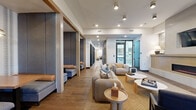 This expansive single-floor amenity space is designed with a modern yet warm aesthetic, blending natural materials with contemporary furnishings throughout. The layout flows from a reception area featuring a light wood desk, a wall-mounted fireplace, and an accent wall with botanical wallpaper, creating an inviting first impression. Recessed and track lighting illuminate the white ceilings, while exposed wood beam accents and large windows allow natural light to filter throughout.

The main lounge zones are furnished with plush, rounded sofas in neutral and amber tones, complemented by organic-shaped coffee tables and woven area rugs. Cozy semi-private booth seating with navy upholstery and pendant lighting offers intimate gathering spots along the corridor. A separate lounge features floor-to-ceiling sheer drapes, a statement chandelier, and tufted leather sectionals.

Dedicated work spaces include glass-enclosed private work-from-home pods and a conference room with a long walnut table and carpeted flooring. Black-framed sliding barn doors with frosted glass panels provide stylish separation between zones. The space connects to a courtyard through glass doors, integrating indoor and outdoor environments seamlessly.