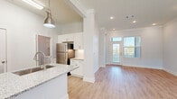 This well-appointed single-floor residence offers a bright, open layout designed for comfortable living. The main living area features light wood-toned flooring, crown molding, recessed lighting, and a glass-paneled entry door flanked by windows that invite abundant natural light. Crown molding runs throughout the main living spaces, adding a refined architectural touch.

The kitchen is a standout feature, centered around a large island with a granite countertop and an integrated double sink. White shaker-style cabinetry lines the walls, complemented by a classic subway tile backsplash, granite countertops, and stainless steel appliances including a full-size refrigerator. Pendant lighting above the island provides both task illumination and visual appeal.

The bedrooms are carpeted for comfort, each offering generous proportions. One bedroom includes a walk-in closet, while another features multiple windows with outdoor views, filling the room with natural light. The bathroom is finished with a white vanity topped with granite, tile wall accents, and wood-look flooring, maintaining a consistent, clean aesthetic throughout.

Additional practical features include a dedicated in-unit laundry space with a top-loading washer. The overall palette of soft whites and light neutrals creates a clean, modern ambiance throughout the entire space.