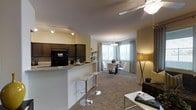 This single-floor residence offers an open-concept layout that seamlessly connects the kitchen, dining, and living areas into one cohesive, functional space. The kitchen features dark cabinetry with a contrasting light granite countertop, black appliances including a refrigerator, and a breakfast bar with modern bar stools — ideal for casual dining or entertaining. Track lighting overhead ensures the kitchen is well-lit and practical. The open layout flows directly into a carpeted dining area, which is anchored by a round dining table with sleek white chairs and illuminated by a decorative chandelier. Large windows dressed with gray curtains allow generous natural light to fill the space, creating a bright and welcoming atmosphere. The living area, situated to the right, is accented by a ceiling fan and a warm floor lamp, contributing to a relaxed and comfortable ambiance. Warm yellow-toned walls throughout lend the space a cozy, inviting feel, while the neutral carpet provides a soft foundation across the main living zones. The overall design balances modern fixtures with warm, residential tones, making the space both stylish and livable.