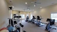 This fitness room offers a dedicated space for exercise and wellness, thoughtfully designed to accommodate a range of workout needs. The room features a neutral color palette with warm beige walls and light carpeted flooring, creating a clean and inviting atmosphere for physical activity.

The space is outfitted with multiple cardio machines, including treadmills and elliptical trainers, arranged in an organized row along one side of the room. A multi-function strength training machine is positioned toward the front, providing options for resistance and weight-based exercises. The layout allows for comfortable movement between equipment without feeling cramped.

Overhead, multiple ceiling fans are evenly distributed throughout the room, ensuring consistent airflow during workouts. Recessed lighting is integrated into the ceiling, providing bright, even illumination across the entire space. Two windows along the far wall allow natural light to enter, enhancing the open and airy feel of the room. A wall-mounted unit, likely a climate control device, is also visible, contributing to year-round comfort. The overall design is functional, straightforward, and well-suited for regular fitness use.