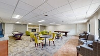 This single-floor amenity space is thoughtfully designed to accommodate a variety of social and recreational activities. The expansive main lounge area serves as the centerpiece, featuring a striking stacked-stone fireplace framed by a wide hearth that anchors the room with warmth and visual character. Surrounding the fireplace are comfortable seating arrangements, including cream leather armchairs and chartreuse green sofas paired with a round ottoman, creating an inviting gathering spot.

The lounge extends into a casual dining and game zone, furnished with round tables and vibrant yellow-green chairs ideal for socializing. A pool table and shuffleboard table add recreational options to the space. Recessed grid ceilings with flush lighting panels illuminate the area uniformly throughout.

A dedicated window seating nook along one wall features large sliding windows that overlook lush greenery and a parking area, filling the space with natural light. Adjacent to this area is a compact kitchenette equipped with light wood cabinetry, granite countertops, black appliances including a range and microwave, and a stainless steel sink. A separate coffee bar station with warm wood cabinetry and granite countertops provides a convenient self-serve refreshment area. Tile flooring in the entry zones transitions to patterned carpet throughout the main lounge.