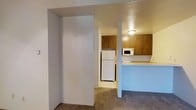 This single-floor studio unit offers a practical and efficiently designed layout that maximizes its compact footprint. The kitchen is a standout feature, equipped with warm-toned wood cabinetry providing ample storage, a full-size refrigerator, a microwave, and a light-colored countertop that wraps around to form a convenient breakfast bar or pass-through divider. The kitchen is separated from the main living area by a structural column and a partial wall, creating a subtle sense of definition between spaces without fully enclosing them. Overhead, a flush-mount ceiling light provides direct illumination to the kitchen area, complemented by a textured ceiling finish throughout. The main living area is carpeted in a neutral tone, lending warmth underfoot and contributing to a cohesive interior palette. A recessed nook or alcove along one wall offers additional spatial versatility, potentially serving as a closet or dedicated storage area. The overall design is straightforward and functional, suited for an individual seeking a low-maintenance, self-contained living space.