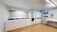 This single-floor communal laundry facility at 872 Bettina Ct in Houston, Texas offers a practical and functional layout designed to serve shared-use needs efficiently. The space features durable, light-toned tile flooring throughout, complemented by neutral gray walls and bright overhead fluorescent lighting that ensures excellent visibility across the entire room.

The laundry area is equipped with multiple commercial-grade top-loading washing machines and a set of stacked dryers, all from Speed Queen, providing ample capacity for regular use. Informational signage and laundry tips are posted on the walls for user convenience. A dedicated folding table with a clean white surface and metal legs offers a practical workspace for handling clean laundry.

A standout feature of the room is the full-size Coca-Cola vending machine, providing easy refreshment access. The space is accessible through white French doors with glass panes, allowing natural light to filter in and offering a connection to the exterior. A wall-mounted bulletin board provides additional informational resources for users.

The room maintains a clean, utilitarian aesthetic with white baseboards framing the flooring and HVAC vents ensuring proper ventilation. Overall, the space is thoughtfully arranged to maximize usability within its modest footprint, making it a convenient and well-appointed shared laundry facility.