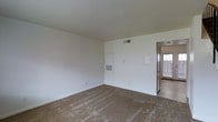 This two-floor residential space presents a straightforward and functional layout designed for comfortable everyday living. The interiors are finished with neutral white walls and textured ceilings, creating a bright and open atmosphere throughout. The main living area features wall-to-wall carpeting in a warm beige tone, providing a soft underfoot feel and a cozy ambiance. Practical built-in utility elements are visible along the walls, including ventilation grilles and access panels, reflecting a thoughtfully designed infrastructure. A doorway leads to an adjacent area that transitions to hard flooring, hinting at a multi-surface layout across the space. Natural light filters in through a window visible near the entryway, adding warmth to the otherwise clean and minimal interior. The white walls and simple trim work offer a versatile backdrop suitable for a variety of interior styles. Spanning two floors, the space provides ample room for distinct living and private zones, making it well-suited for those seeking a functional, no-frills layout with room to personalize. The overall aesthetic is clean, neutral, and move-in ready.