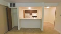 This single-floor space is designed with an open, functional layout that promotes a seamless flow between the kitchen and living areas. The kitchen is framed by a distinctive pass-through cutout in the wall, creating a visual connection to the adjoining carpeted living space while maintaining a defined cooking zone. White countertops extend along the pass-through, offering a casual bar-style surface ideal for dining or additional prep space.

Inside the kitchen, warm medium-toned wood cabinetry lines the upper walls, providing ample storage. Essential appliances, including a white refrigerator and a gas range with overhead ventilation, are neatly integrated into the compact layout. Recessed flush-mount ceiling lights illuminate the kitchen evenly, contributing to its clean, neutral aesthetic.

The living area features wall-to-wall carpet in a neutral tone, enhancing the warmth of the space. White walls and ceiling throughout maintain a bright, cohesive appearance. A closet is partially visible along one wall, adding practical storage options. The overall ambiance is clean, understated, and move-in ready.
