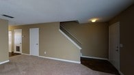 This two-floor space is defined by a practical and straightforward layout. The main level showcases a transition between two distinct flooring surfaces — a carpeted area and a section of dark hardwood flooring — offering visual contrast throughout the space. Warm, tan-toned walls are complemented by crisp white baseboards and trim, lending the interior a clean, cohesive appearance. A textured popcorn ceiling adds a classic touch overhead.

A centrally positioned staircase with white-painted railings and trim connects the two floors, serving as a functional architectural focal point within the room. The staircase rises along the back wall, with the upper level partially visible from below. A flush-mount ceiling light near the staircase provides illumination to the area. A single white door on the main level provides access to an additional room or closet space. Electrical outlets are visible along the baseboards, and the overall layout suggests an open, livable lower level that flows toward the stairwell leading to the upper floor.