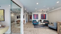 This single-floor space serves as a welcoming lobby or reception area, designed with a modern aesthetic that balances functionality and style. The entry is framed by a glass door etched with a "V" logo and hours of operation, immediately establishing a professional yet approachable atmosphere.

The flooring is a standout feature, showcasing light-toned wood laid in a distinctive herringbone pattern that adds visual depth and warmth throughout the space. Recessed lighting is evenly distributed across the white ceiling, supplemented by a sculptural, Edison-bulb chandelier that introduces an artistic focal point and soft ambient glow.

The seating area is anchored by a pair of teal mid-century modern armchairs and a matching sofa arranged around a round white coffee table, all set atop a patterned area rug. Two large, bold abstract paintings in red and blue tones dominate the wall, adding vibrant color contrast to the otherwise neutral palette.

Toward the rear, elevator doors and a corridor suggest access to additional floors or areas beyond, contributing to the sense of a functional, multi-use building entrance. Overall, the space conveys a curated, contemporary ambiance that feels both inviting and polished.