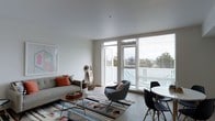 This thoughtfully designed single-floor space offers a clean, contemporary aesthetic characterized by an open-plan layout that connects the living and dining areas in a cohesive, functional arrangement. The living area is anchored by a comfortable sofa accented with warm terracotta throw pillows, paired with a sculptural accent chair and a low-profile coffee table resting atop a geometric, multi-colored area rug that adds texture and visual interest to the space. A framed abstract artwork on the wall contributes a touch of artistic character. Large floor-to-ceiling windows flood the interior with abundant natural light, while horizontal blinds offer adjustable privacy and light control. A glass door opens directly to a private balcony, extending the living space outdoors and offering pleasant views of surrounding greenery and the neighborhood beyond. The dining area is neatly positioned adjacent to the living space and features a round white table with sleek black modern chairs, creating an intimate yet stylish setting for dining. The smooth white ceilings and neutral toned walls and flooring throughout maintain a light, airy ambiance that enhances the sense of openness. Overall, the space strikes a balance between modern design sensibility and comfortable everyday living.