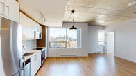 Located in the heart of downtown Montréal on Rue De Bleury, this well-appointed 2-bedroom, 2-bathroom condo offers a thoughtfully designed layout with contemporary finishes throughout. The open-concept living and kitchen area serves as the heart of the unit, where light hardwood flooring and an exposed concrete ceiling lend an industrial-modern character. The kitchen is outfitted with sleek white high-gloss cabinetry, a white tile backsplash, stainless steel appliances including a refrigerator, stove, built-in microwave, and dishwasher, all anchored by a bold pendant light fixture. Large windows flood the living area with natural light and frame impressive views of the Montréal skyline.

The primary bedroom is a comfortable retreat featuring floor-to-ceiling sliding glass doors that open to a private balcony with city views, along with a built-in closet. The second bedroom also benefits from generous window exposure and the same warm hardwood flooring. Both bathrooms are finished with large-format floor tiles and warm wood vanities topped with white basins. The main bathroom features a glass-enclosed corner shower with white subway tile, while the second bathroom offers a similarly refined aesthetic. The exposed concrete ceiling continues as a unifying design element throughout the unit.